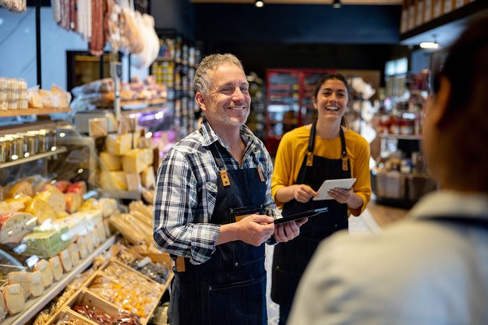 Two deli owners with tablets talking with a customer across the counter