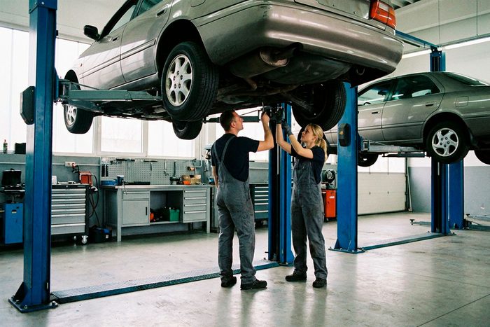 Two technicians work beneath a car raised on a lift in a repair shop, a second vehicle up on the next lift.