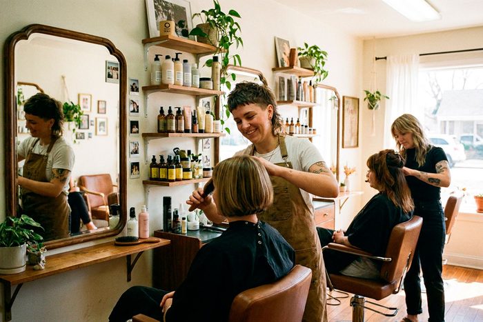A stylist combs out a client’s cut at the chair in a small salon, a second stylist working with another client behind her.